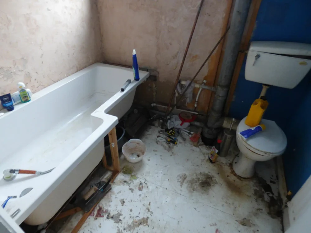 A grubby, dated bathroom with exposed pipework and a stripped floor in a poor condition house