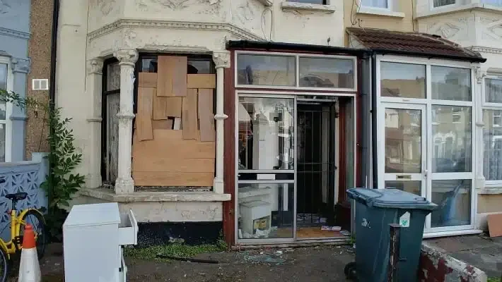 A derelict terraced house with a boarded-up, broken window and rubbish outside