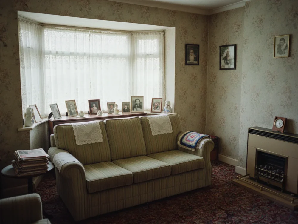 A homely but dated living room in a modest house lived in by a long-term sitting tenant, with patterned wallpaper, net curtains and framed family photographs